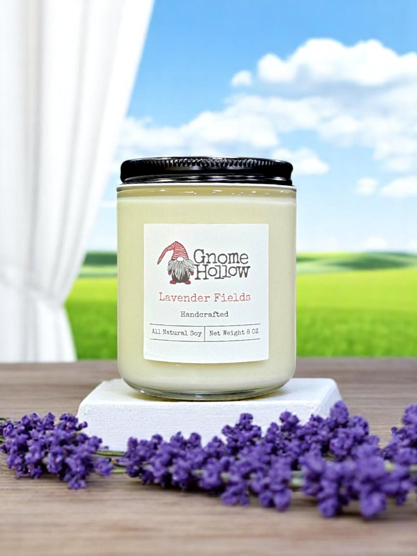 Lavender Fields candle placed on a white surface, surrounded by lavender flowers with a natural landscape in the background.
