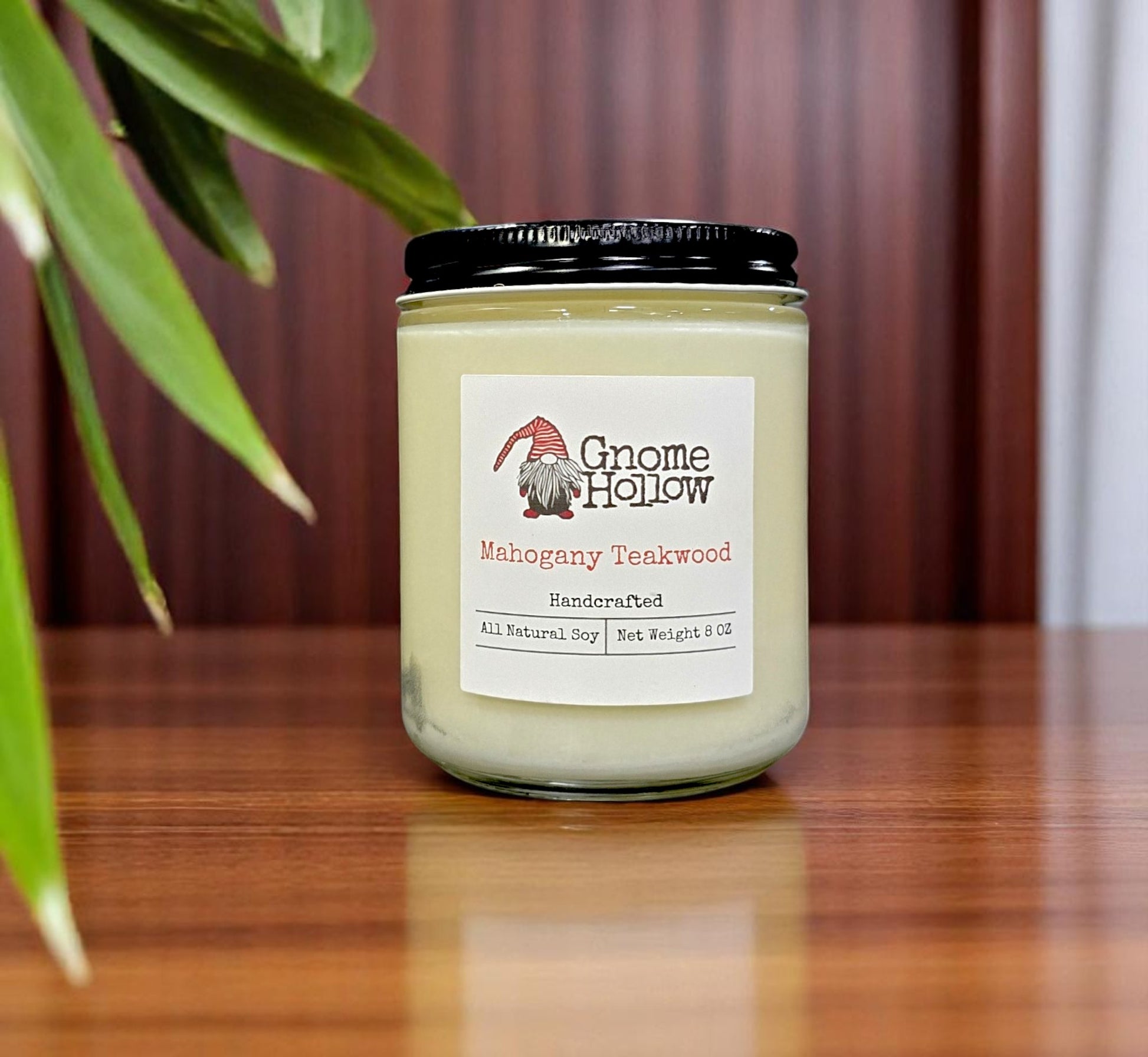 Mahogany Teakwood candle placed on a wooden surface, with a plant leaf in the background.