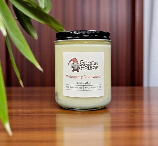 Mahogany Teakwood candle placed on a wooden surface, with a plant leaf in the background.