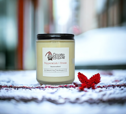 Peppermint + Cream candle sitting in a dusting of snow with a red flower to its right