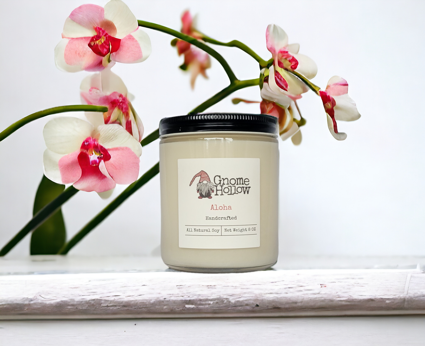 Aloha candle placed on a white surface, with pink and white orchid flowers elegantly framing the background.