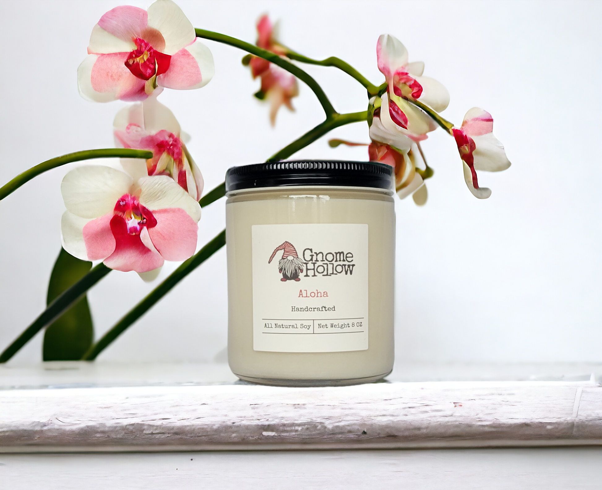 White handcrafted candle in a jar labeled 'Gnome Hollow - Aloha' placed on a white surface, with pink and white orchid flowers elegantly framing the background.