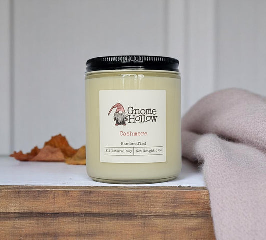 Cashmere candle placed on a wooden surface, with autumn leaves and a pink scarf in the background.