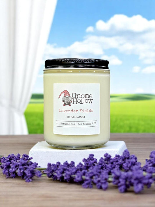 Lavender Fields candle placed on a white surface, surrounded by lavender flowers with a natural landscape in the background.