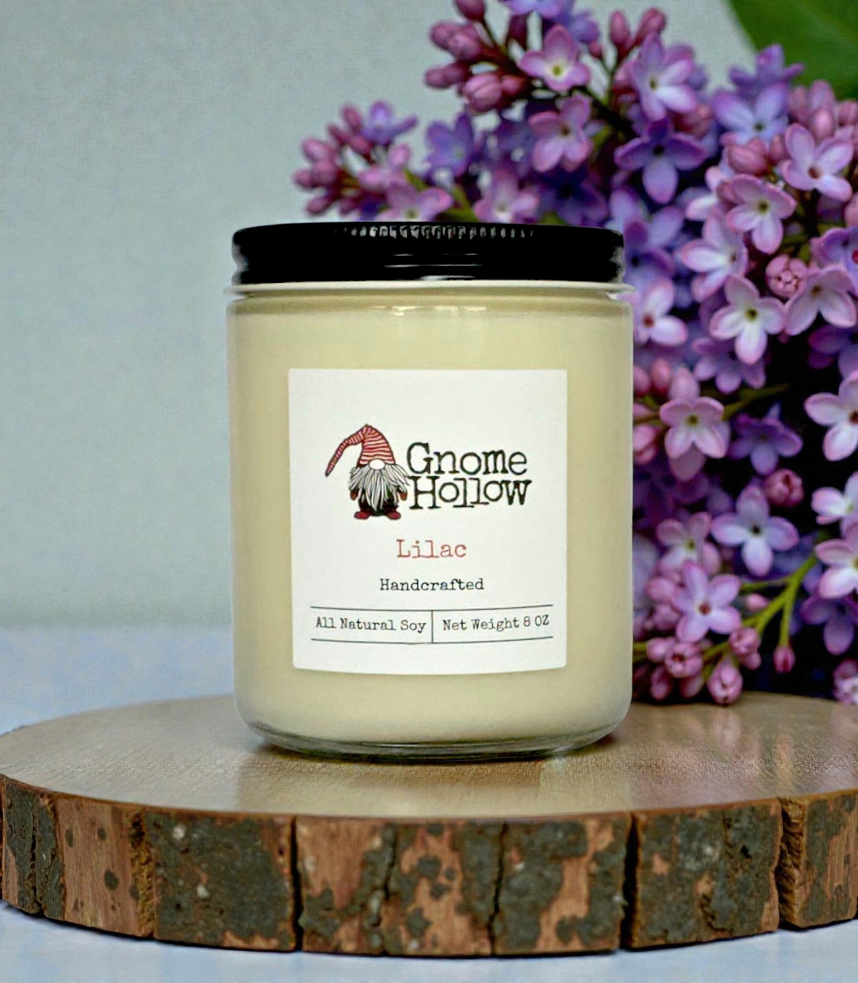 Gnome Hollow Lilac candle on a wooden stand with purple flowers in the background
