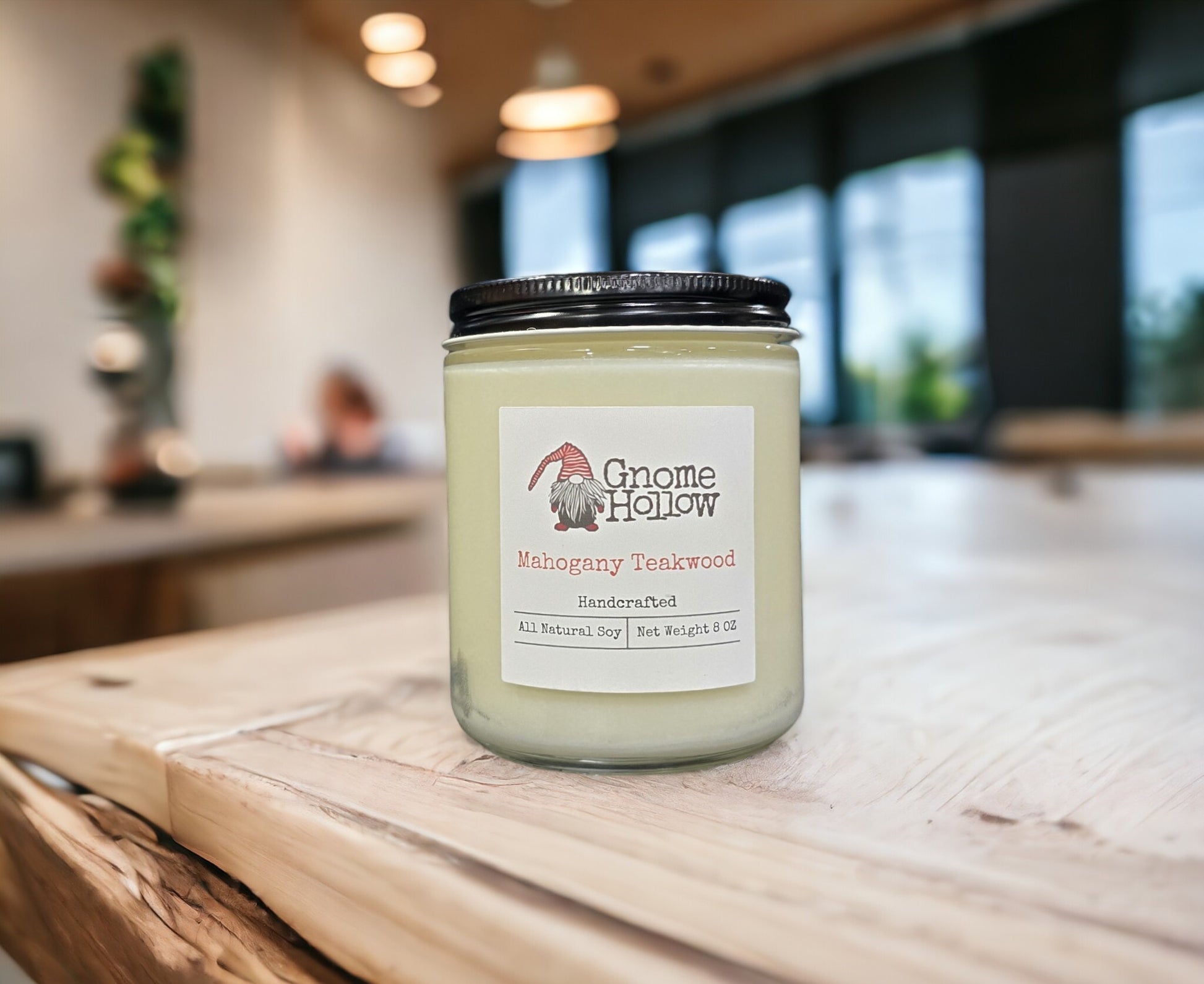 Mahogany Teakwood candle placed on a wooden surface in a room with windows and a plant.