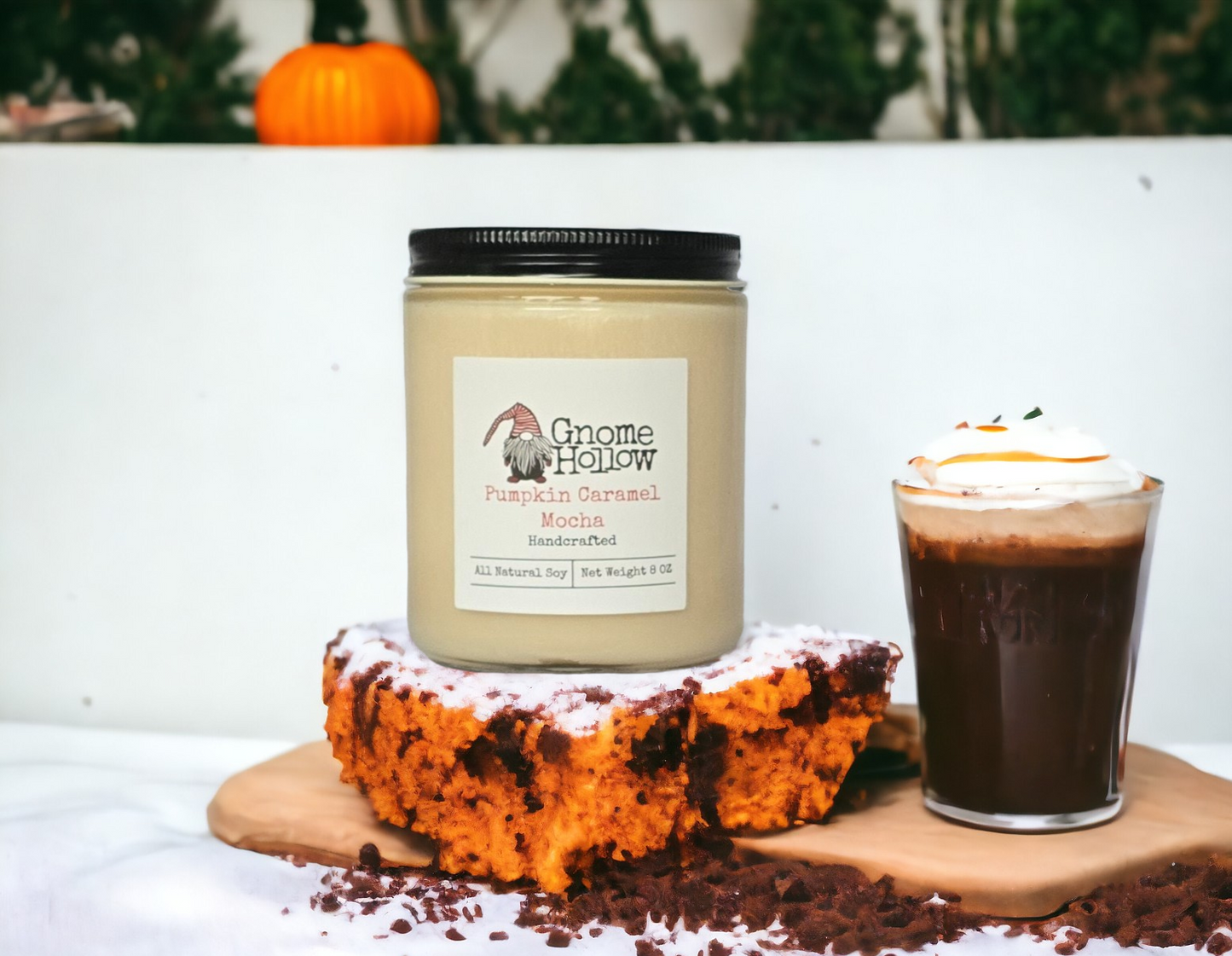 A jar of pumpkin caramel mocha candle with a label that reads 'Gnome Hollow Candles', placed on a wooden surface with a piece of cake and a cup of coffee in the background, suggesting a cozy, autumnal theme.