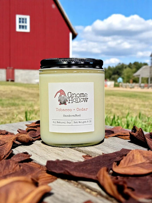 Tobacco + Cedar candle sitting on a picnic table surrounded by tobacco leaves with a red barn in the background