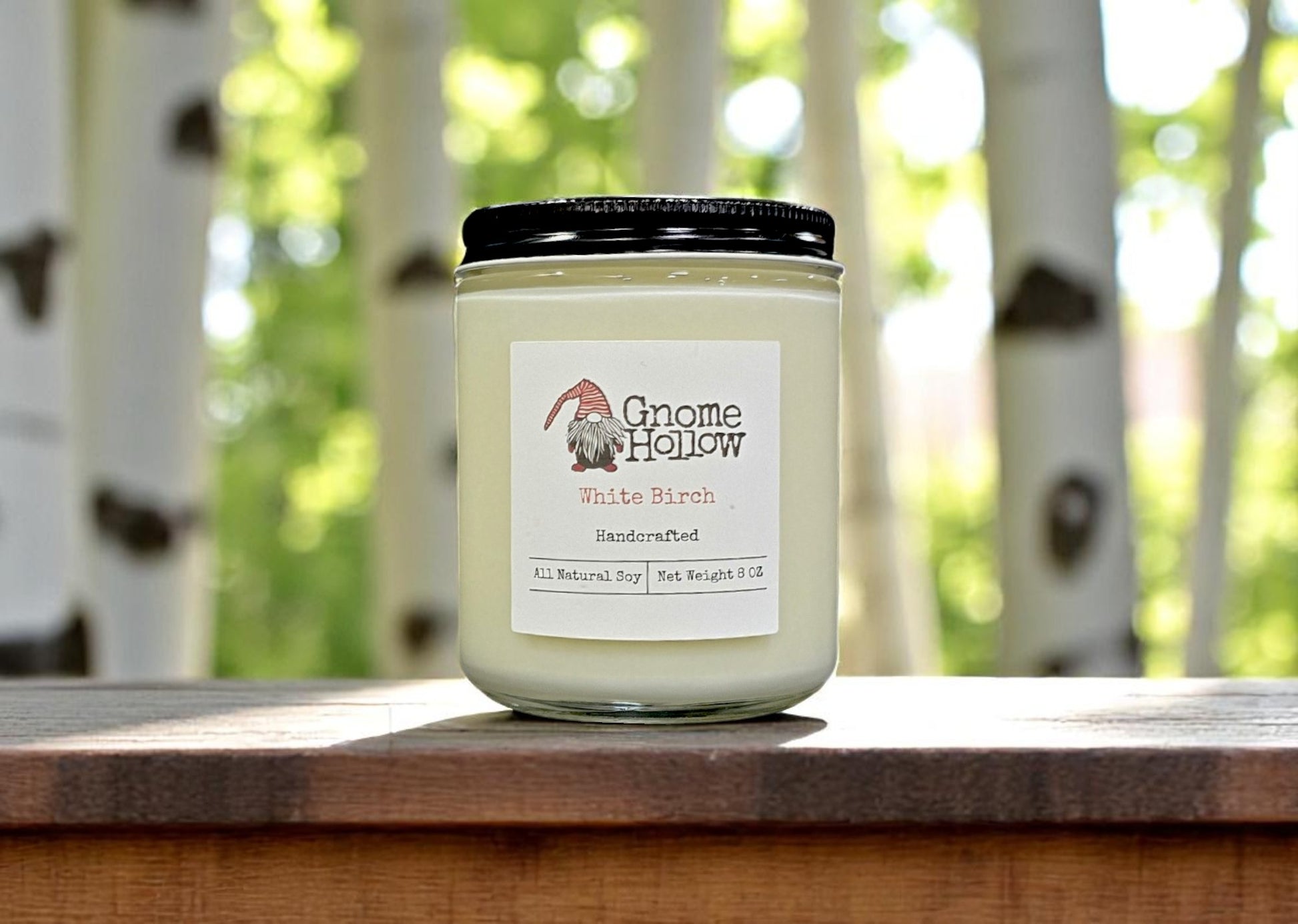White Birch candle sitting on a wood railing with white birch trees in the background